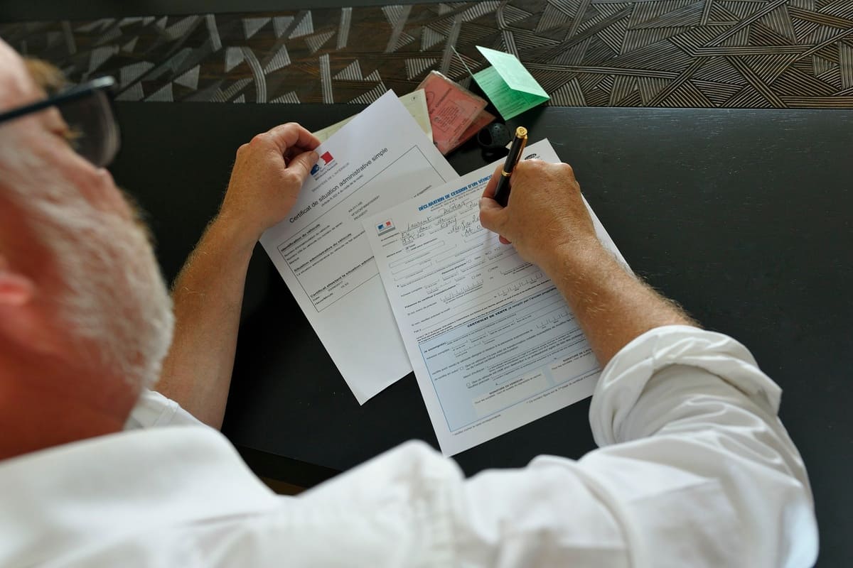 un homme en train de remplir au stylo le formulaire Cerfa 15776 avec le certificat de situation administrative et la carte grise posés sur la table 